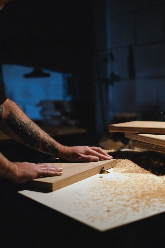 Unrecognizable male artisan trimming edges of wooden board while working at workbench with shavings in professional dark joinery