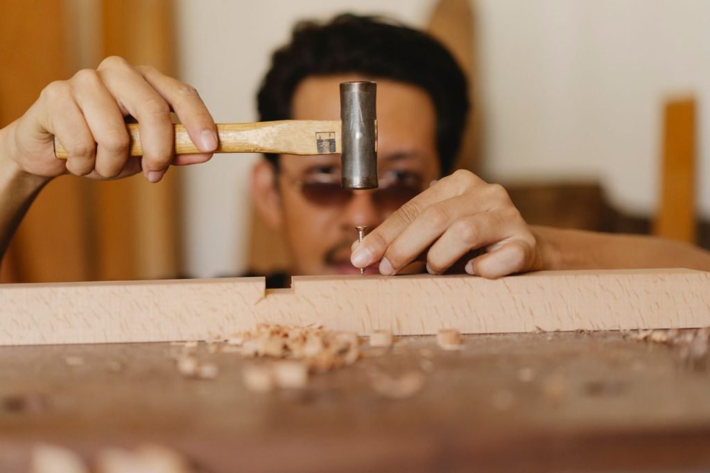 Concentrated young ethnic craftsman in eyeglasses hammering nail into wooden plank during work in carpentry studio